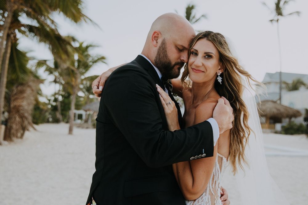 Romantic couple embraces on sandy tropical beach with palm trees swaying in background on sunny day.