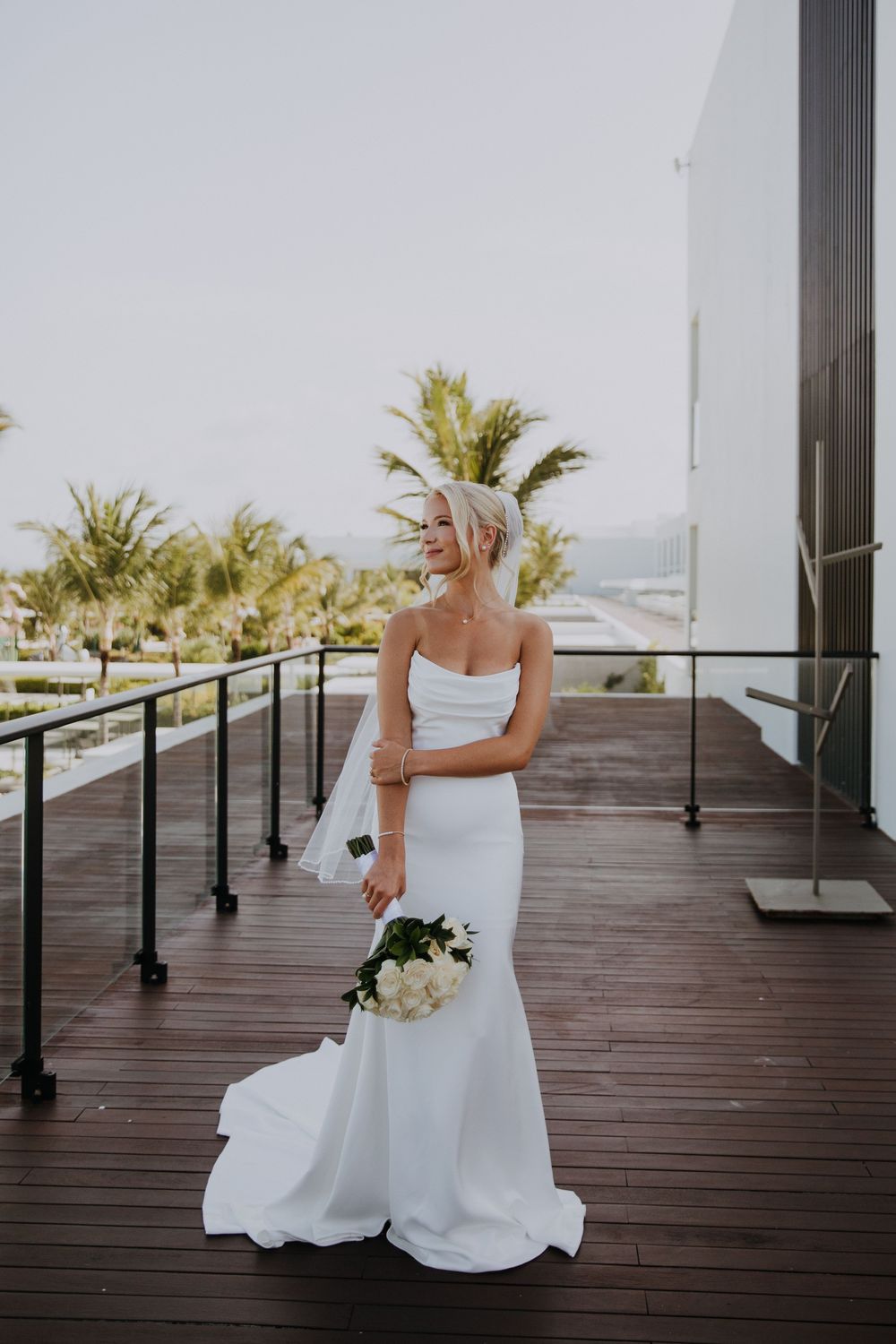 Bride in a white wedding dress sits thoughtfully on a wooden deck overlooking palm trees and ocean views.