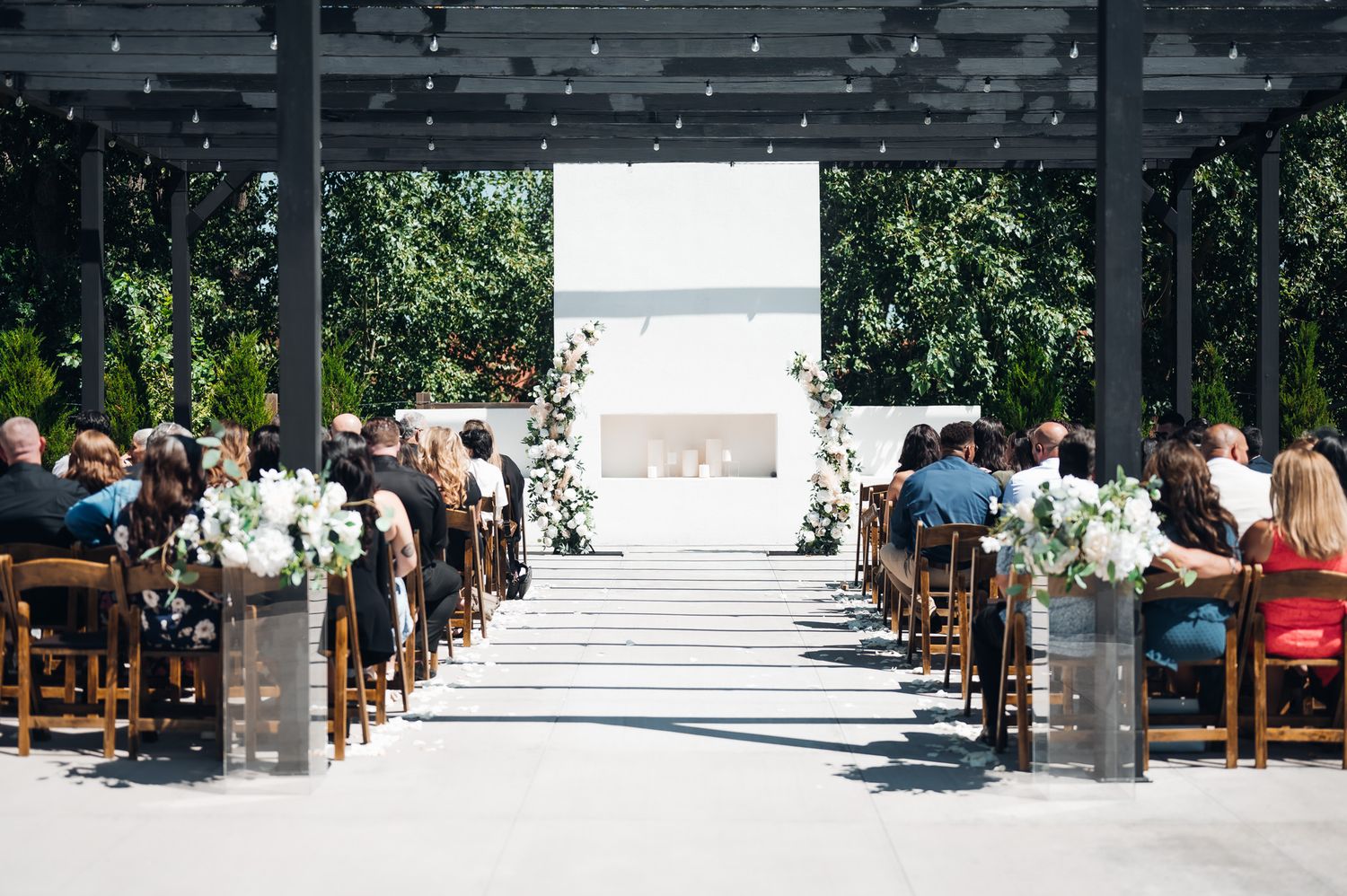 Panoramic view shows stadium-style seating setup with white chairs in an outdoor garden space filled with trees.