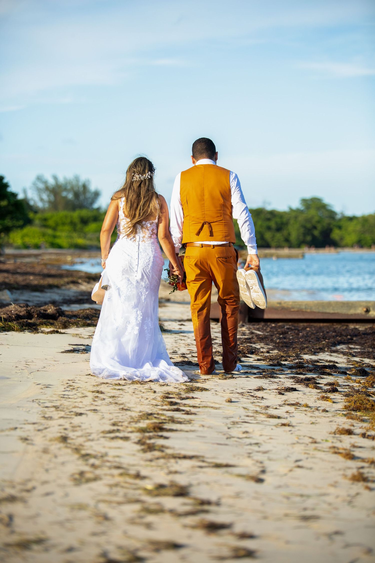 A couple wearing a white dress and orange outfit sit together on a sandy beach near the ocean.