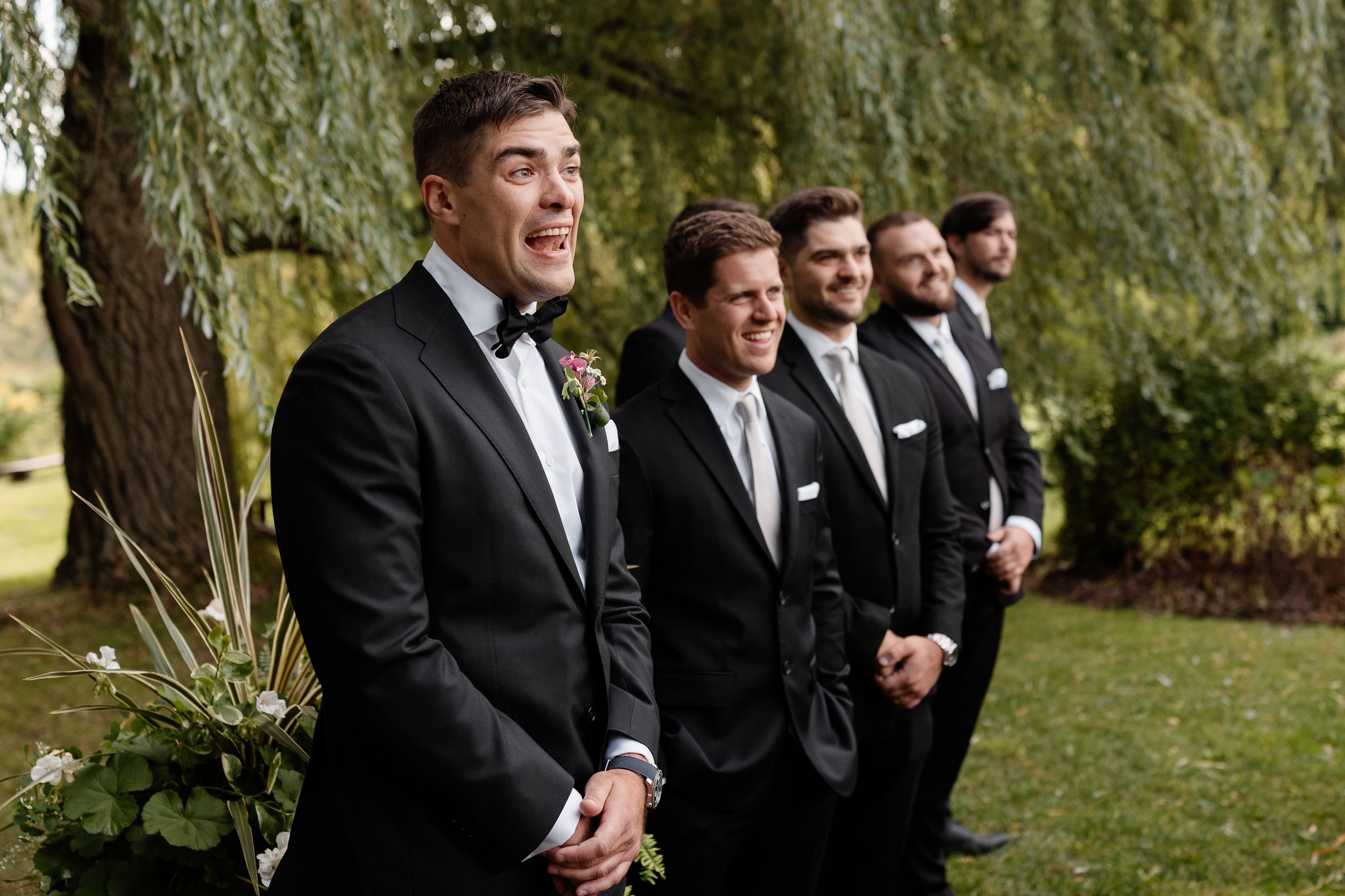A wedding party stands together outdoors under a large weeping willow tree on a sunny day.
