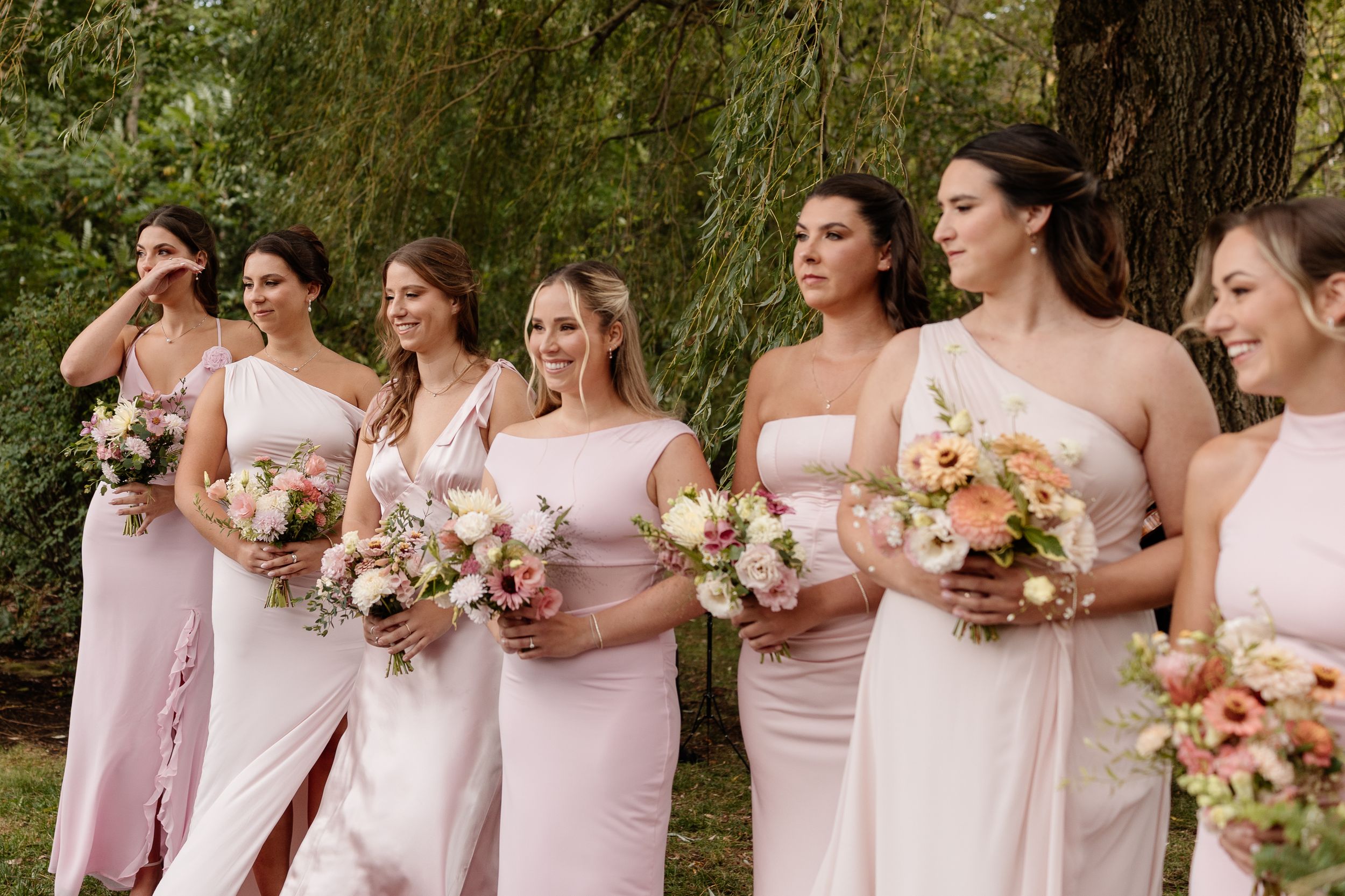 Bridesmaids in blush pink dresses stand together holding peach and white floral bouquets during an outdoor wedding.