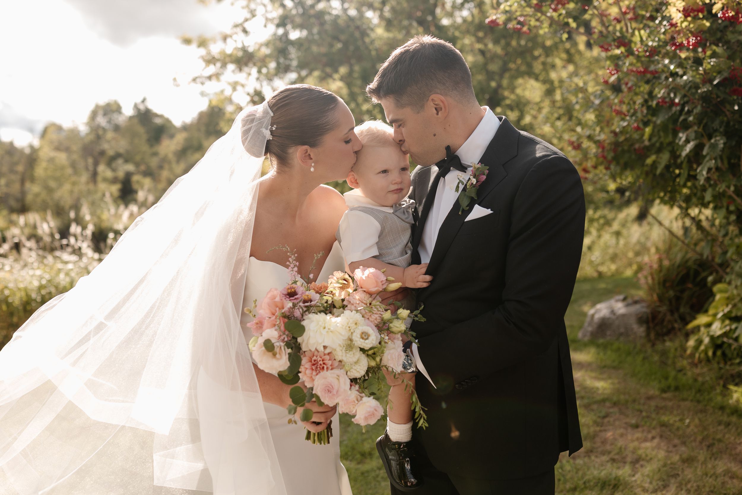 A romantic wedding couple shares tender moments in a garden setting with soft golden sunlight and pink flowers.