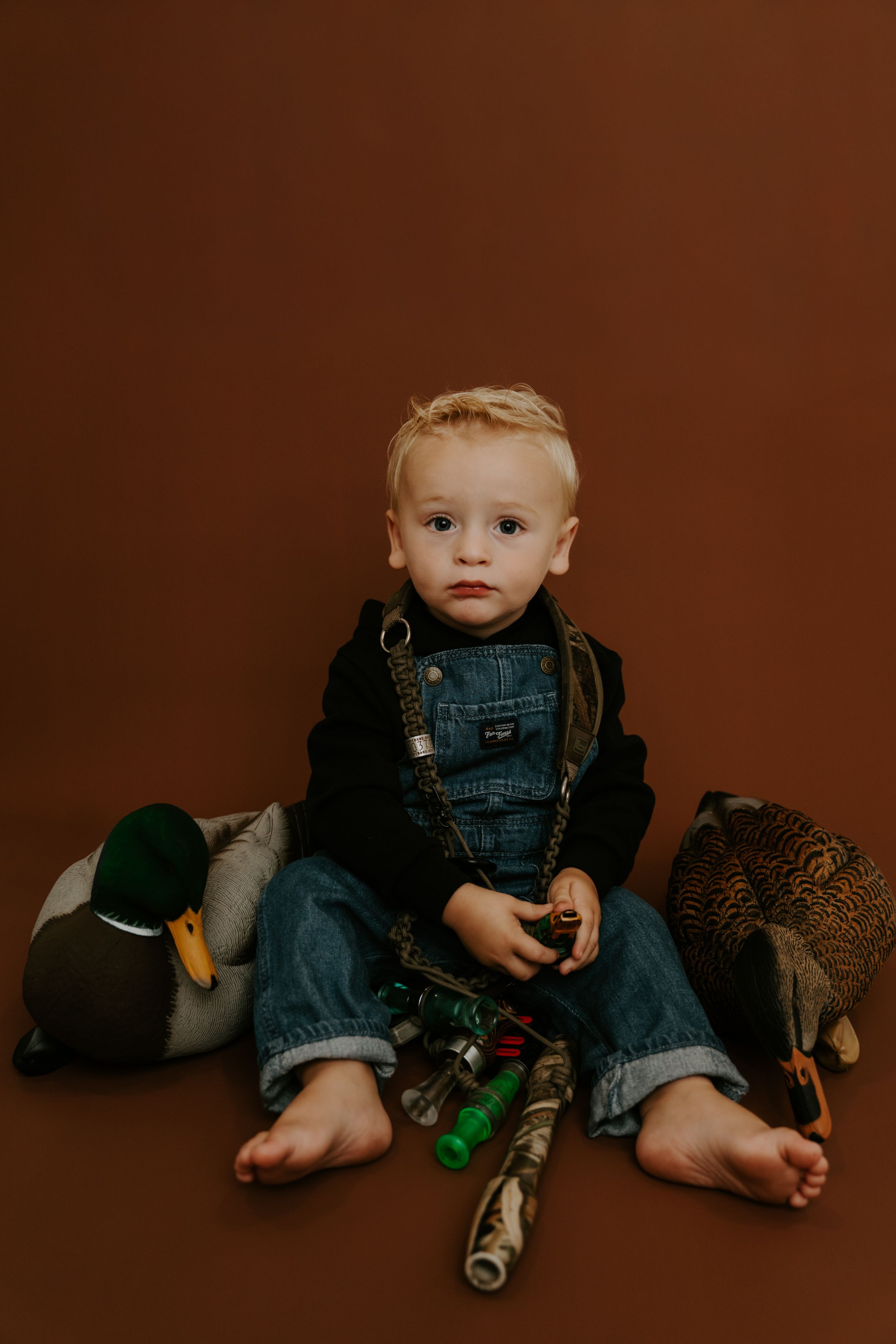 Baby in denim overalls sitting with stuffed animals against dark brown backdrop.