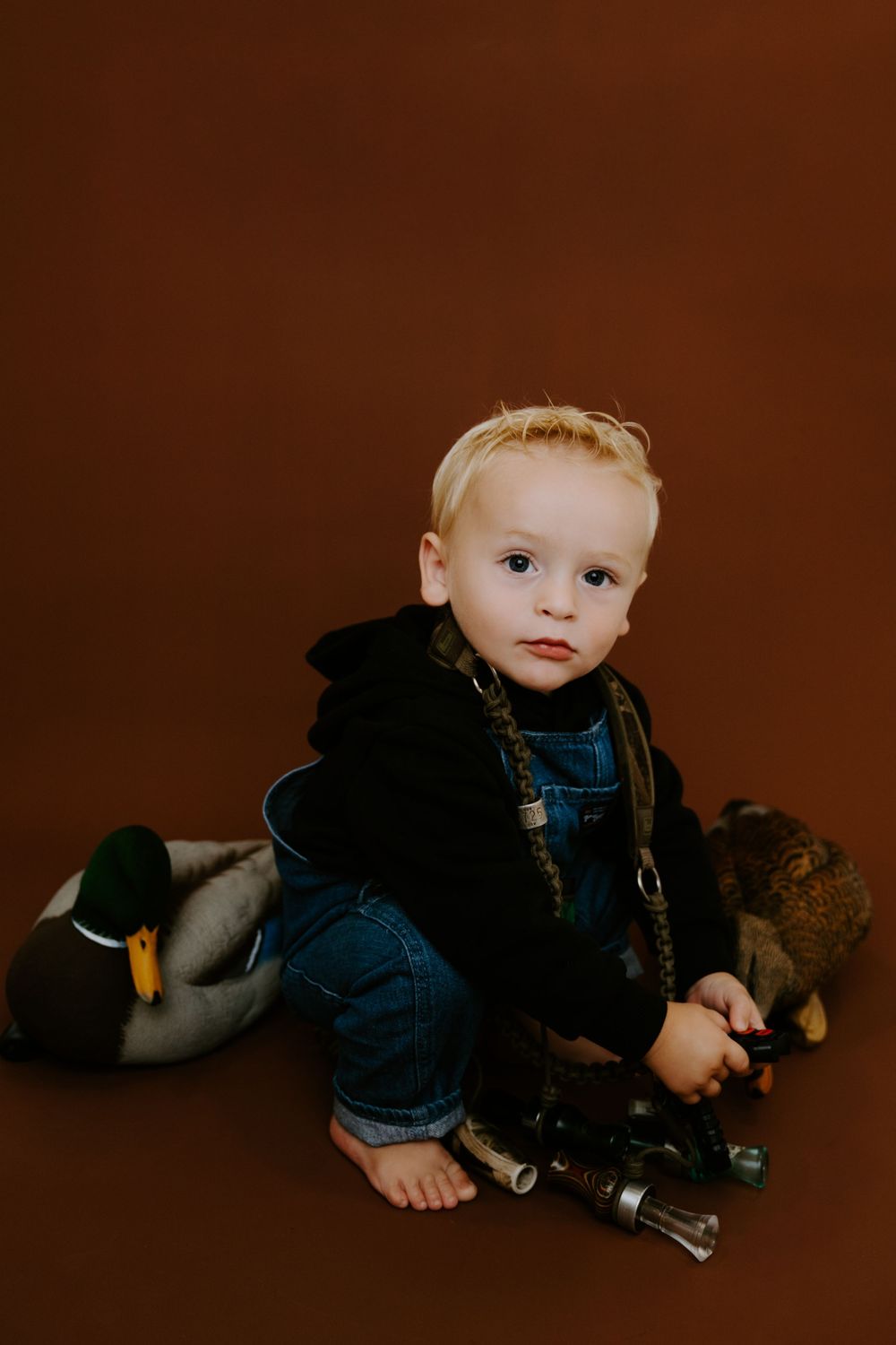 Baby in blue overalls poses with plush toys on chocolate brown background.