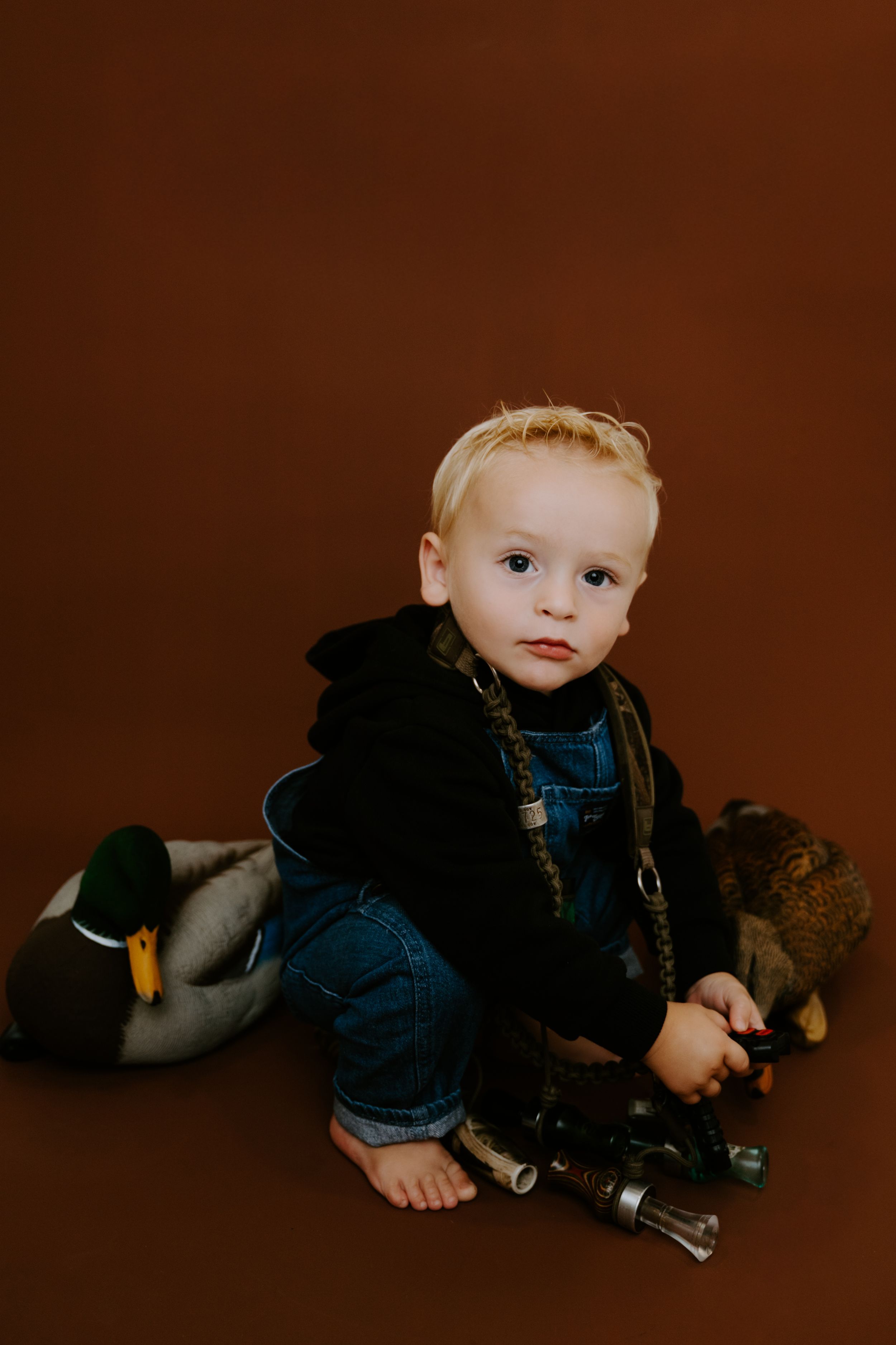 Baby in blue overalls poses with plush toys on chocolate brown background.