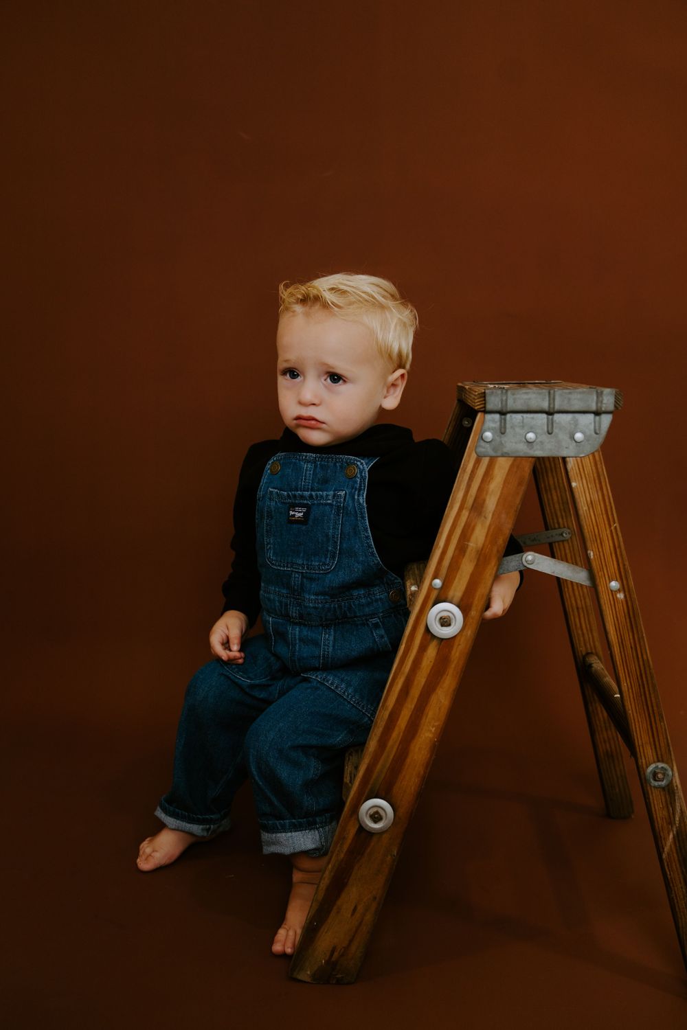 Baby in denim overalls sitting near wooden ladder against brown studio backdrop.