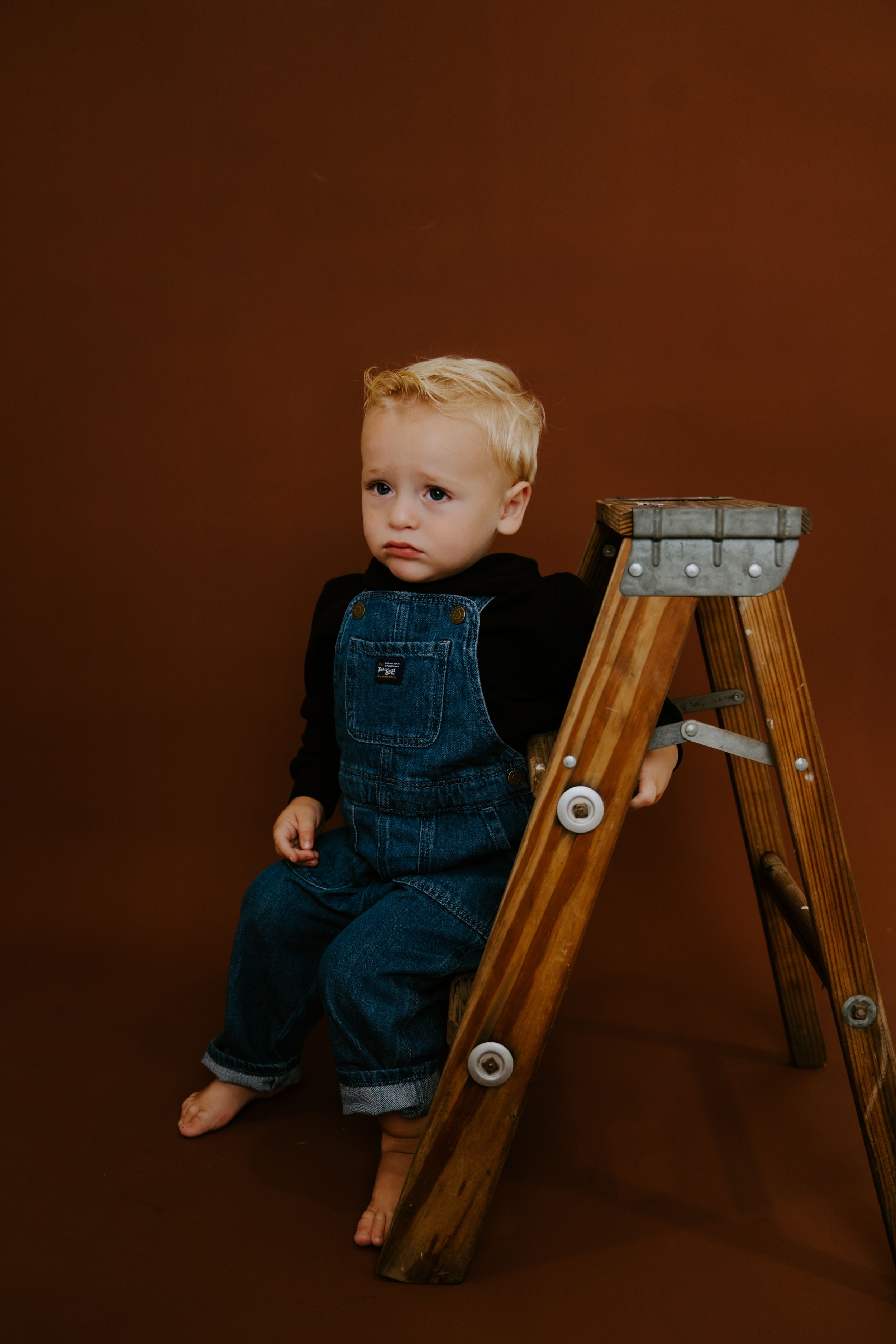 Baby in denim overalls sitting near wooden ladder against brown studio backdrop.