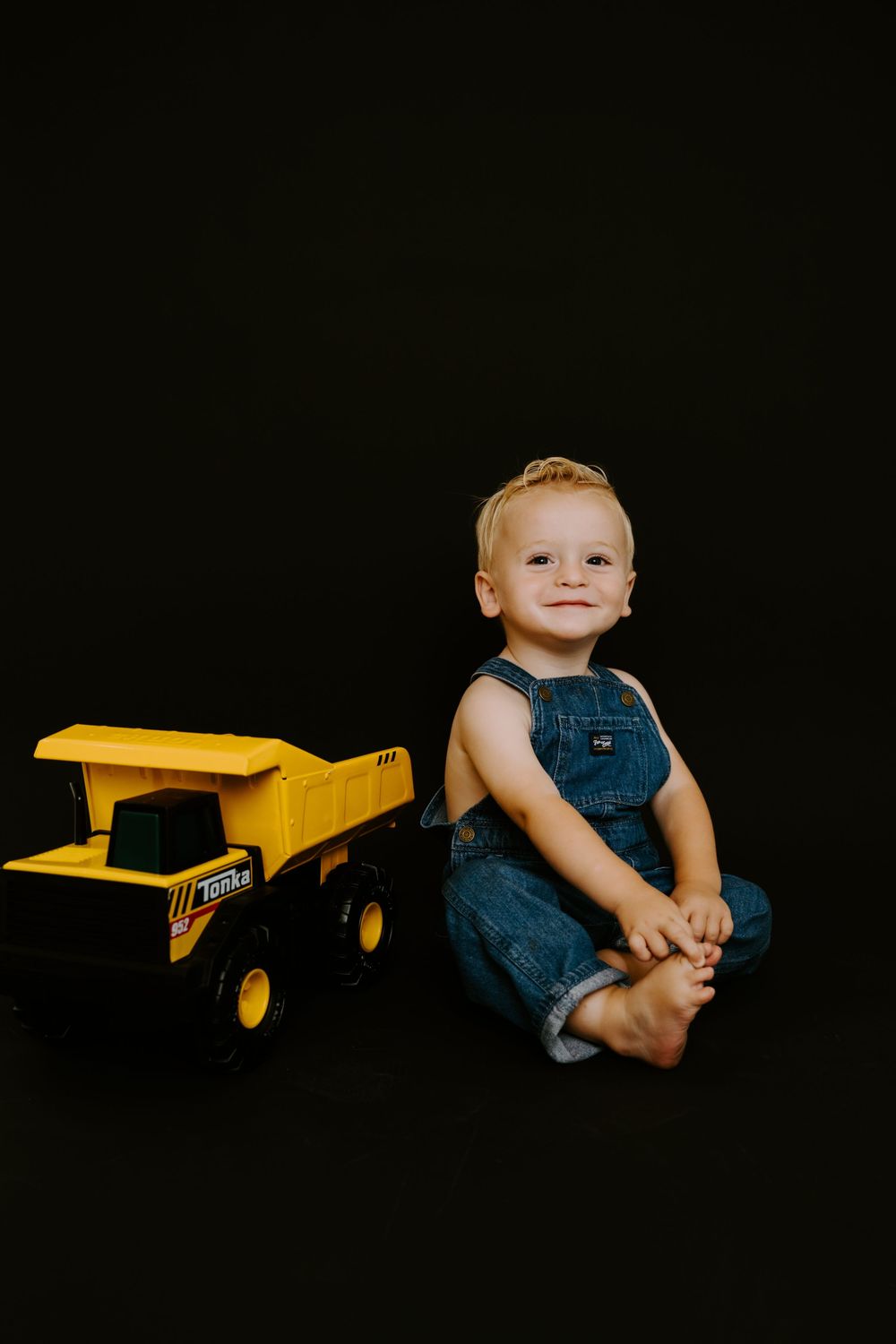 Baby in overalls sitting beside yellow toy truck on black background.