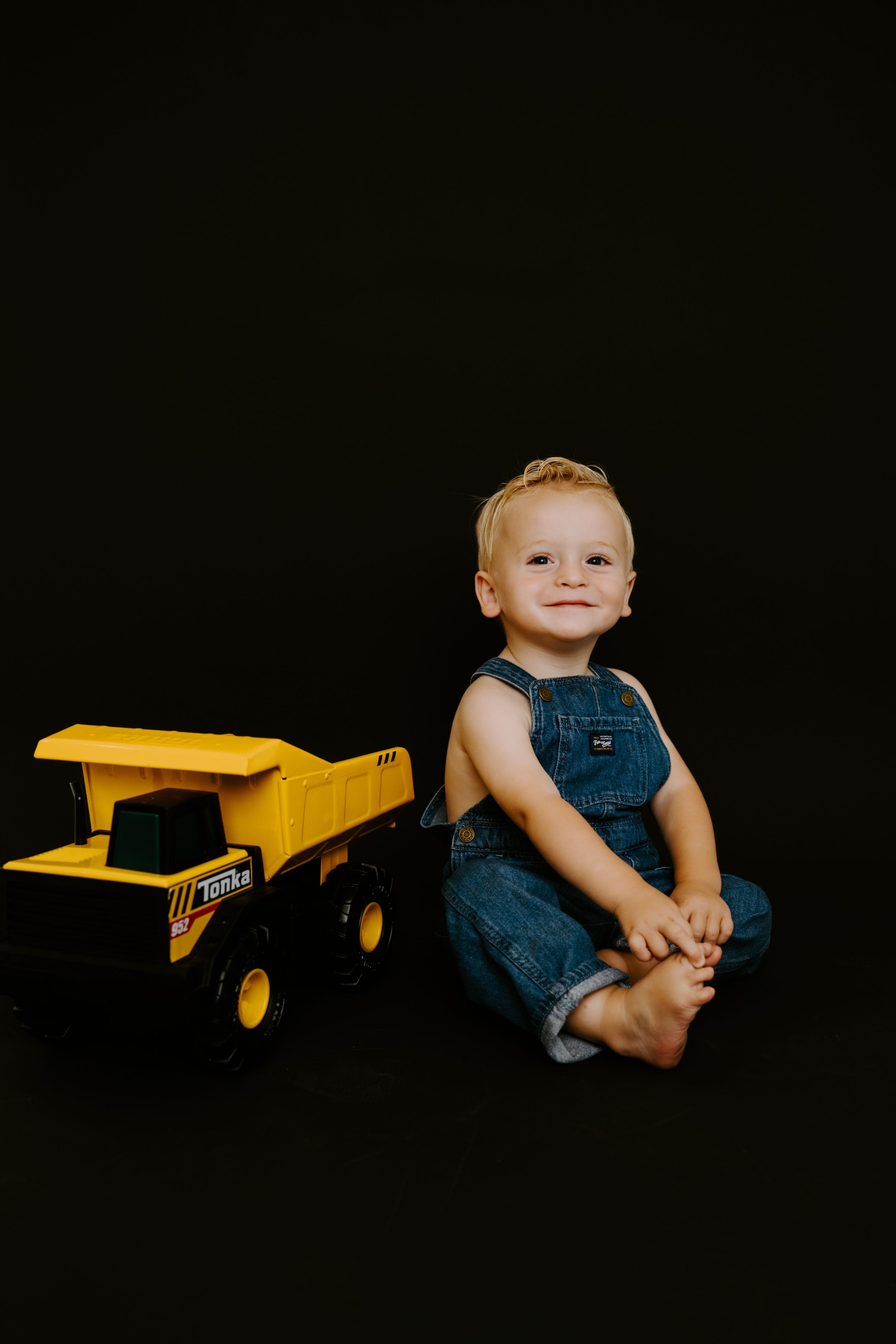 Baby in overalls sitting beside yellow toy truck on black background.