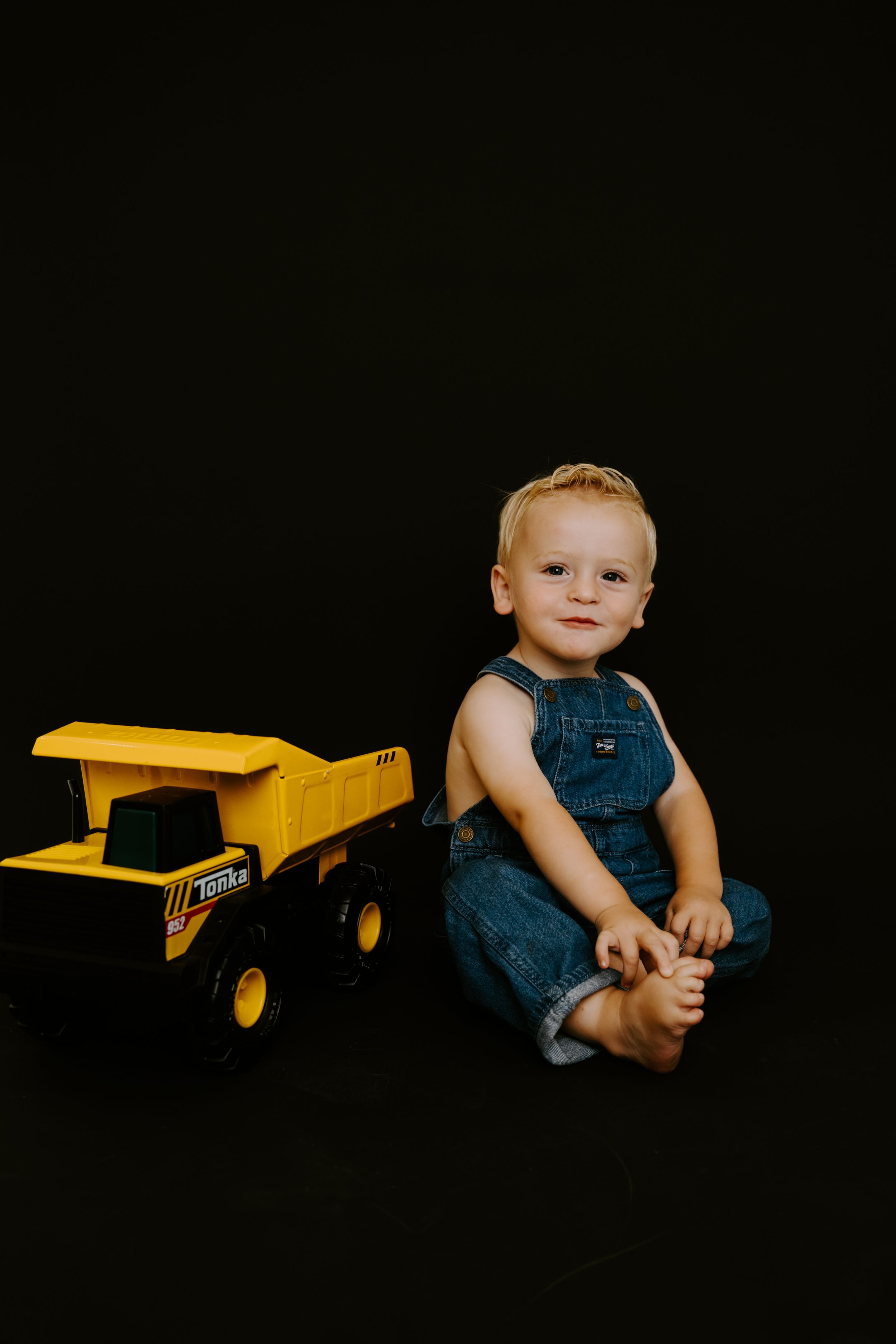 Baby wearing denim overalls poses with yellow toy truck in dark studio setting.