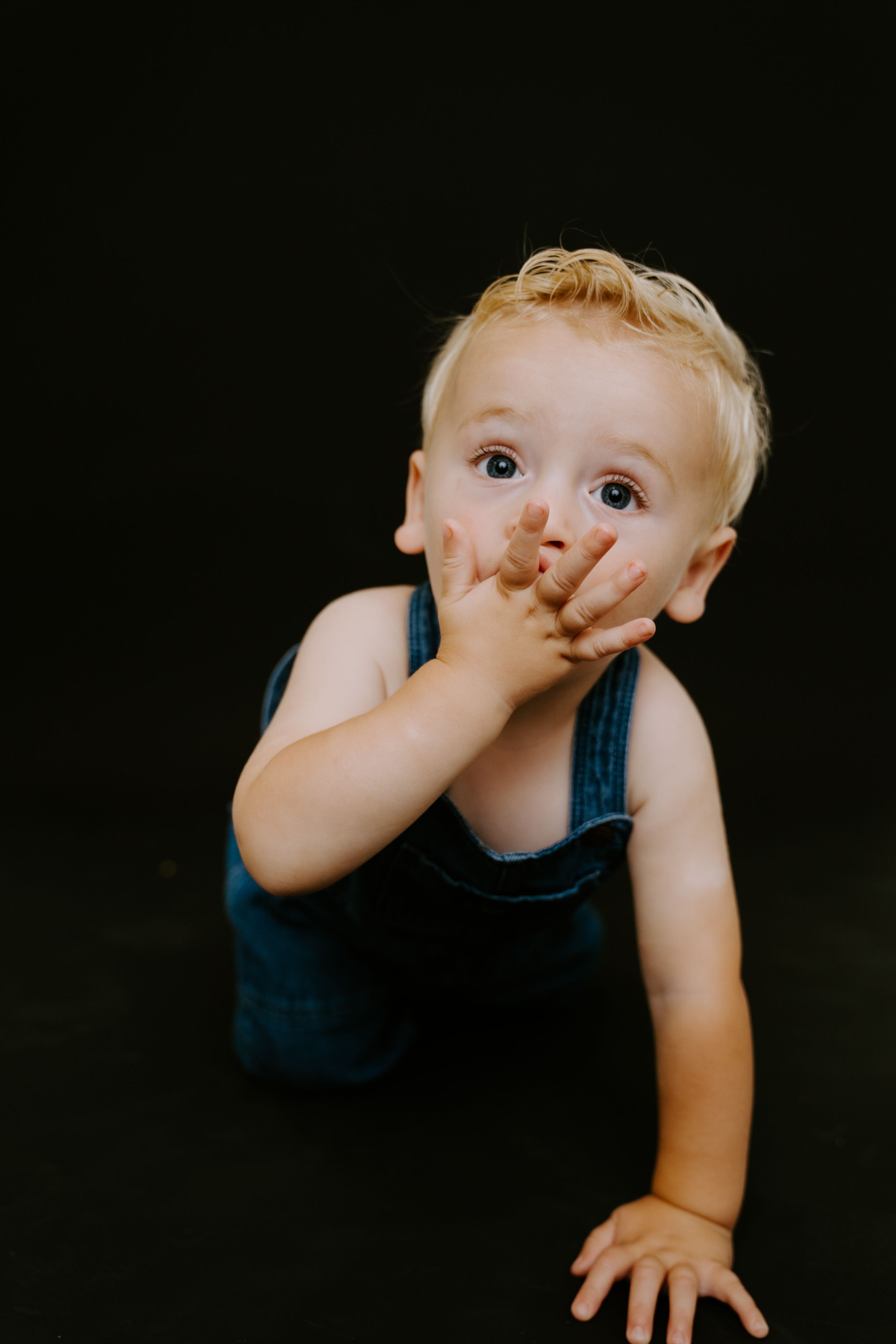 Close-up of a toddler in a black tank top covering their face playfully against a dark background.