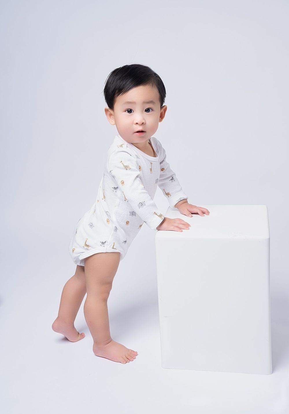A baby wearing a white patterned onesie sits and plays with a white board against a light gray background.