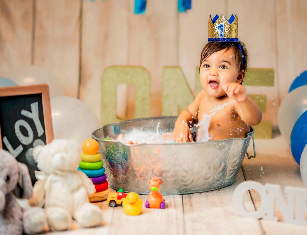 Baby in yellow duck costume enjoying birthday cake smash photoshoot with blue and yellow decorations in studio.