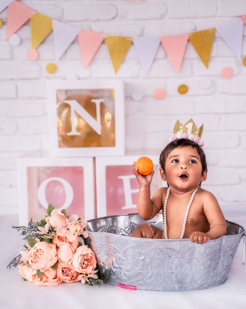 Baby bathtub photo session with peach roses and gold and pink banners against white brick wall background.