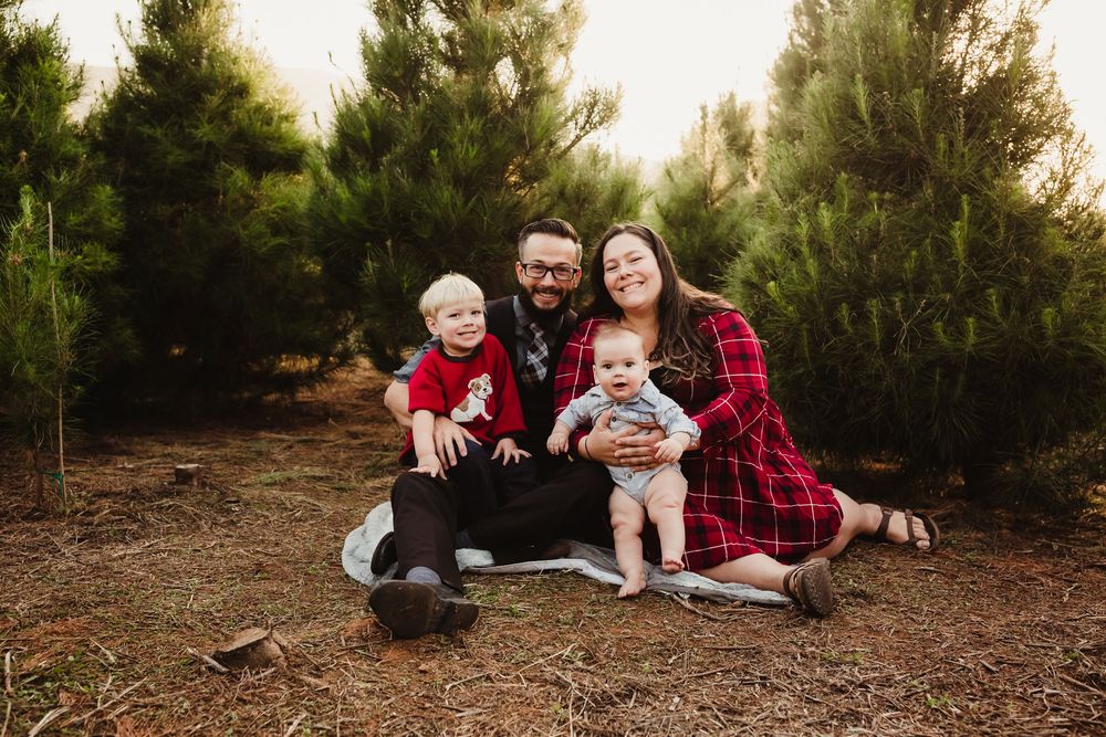 A family wearing red plaid sits together for a photoshoot on a ground covered in pine needles under evergreen trees.