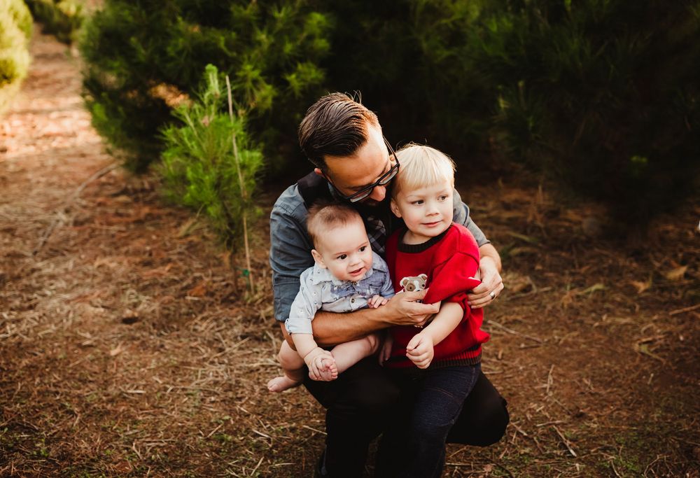 A parent and two small children look at something together while sitting on a dirt path in a wooded area.