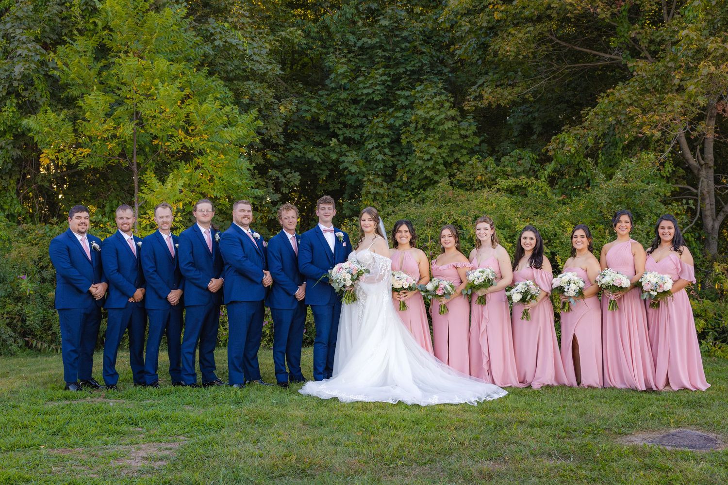 A wedding party in pink and navy blue attire poses for formal photos outdoors in a lush green garden setting.