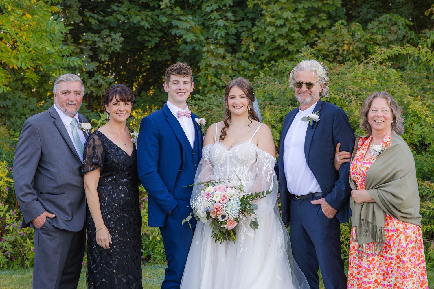 A wedding group poses together outdoors with greenery in the background on a sunny summer day.