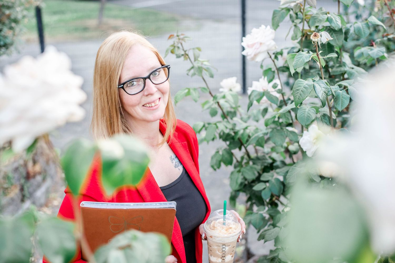 Woman in red blazer smiling among garden plants and white flowers.
