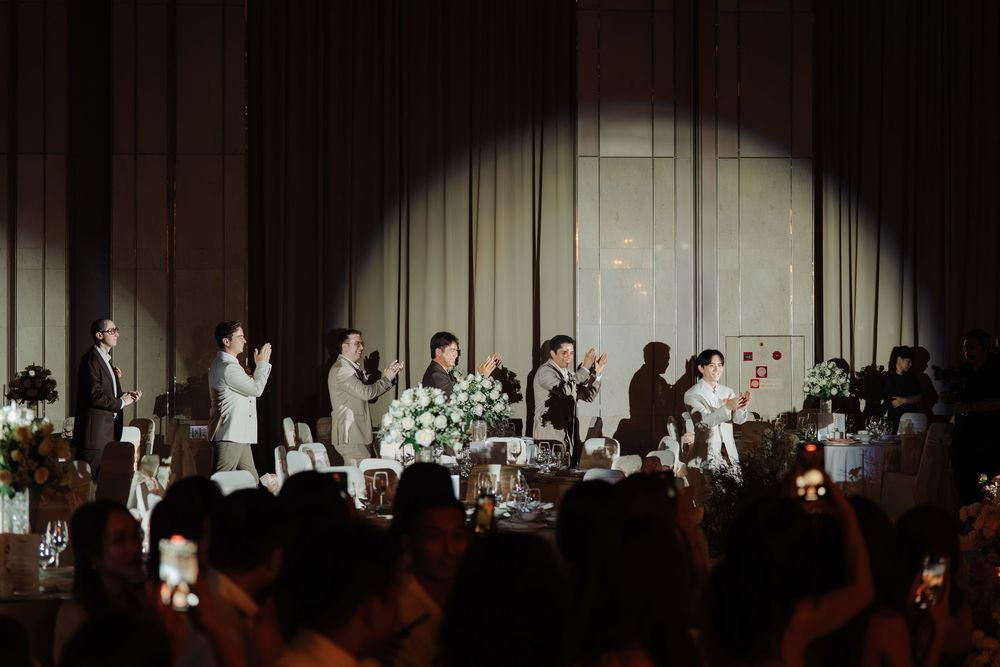 Wedding reception guests gathered on stage under spotlight with floral arrangements.
