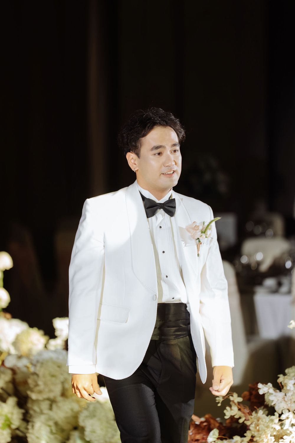 Groom in white tuxedo jacket with black bow tie and boutonniere smiling at wedding reception.