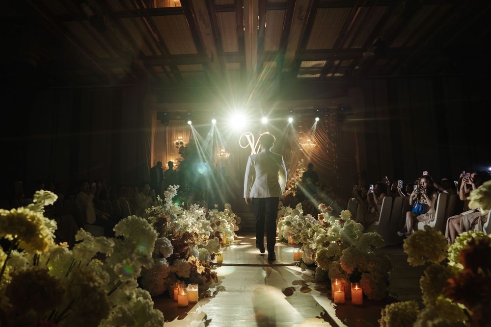 Groom walks down flower-lined aisle toward bright stage lights at romantic wedding ceremony.