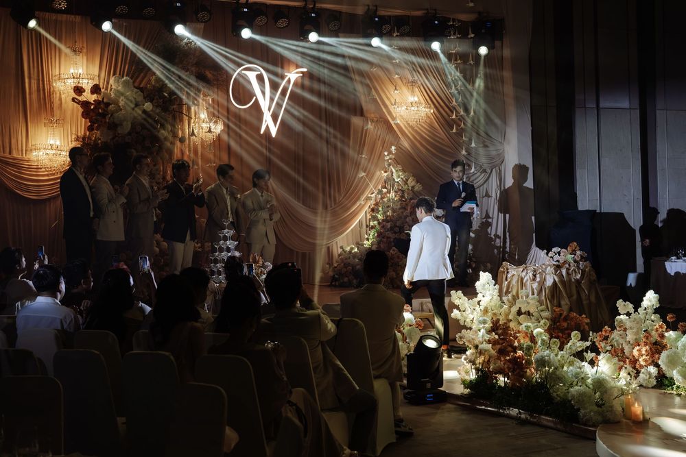 Elegant wedding reception hall with monogram V backdrop, floral arrangements, and guests seated at tables.