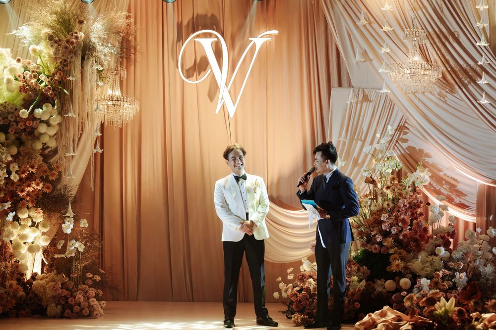 Groom in white jacket stands with MC on stage at wedding reception with monogram V backdrop.