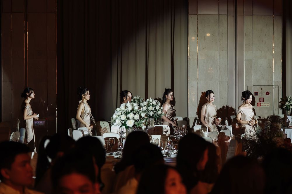 Bridesmaids in champagne gowns walk gracefully past floral centerpieces at wedding reception.