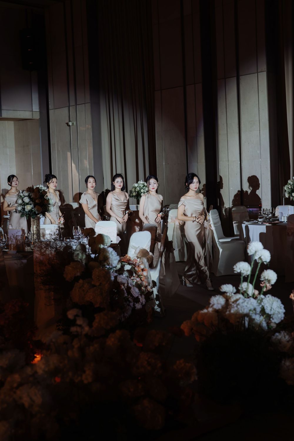 Bridesmaids in elegant champagne dresses seated together at a dimly lit wedding reception.