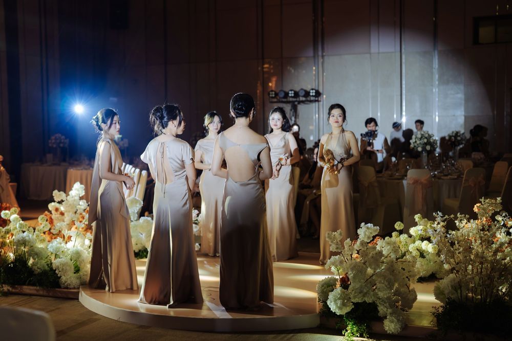 Bridesmaids in champagne dresses gather on illuminated stage surrounded by white floral arrangements.