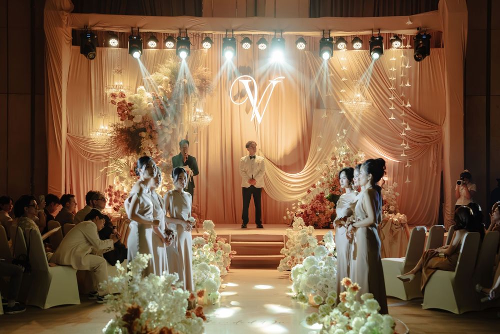 Wedding ceremony with bride and groom at floral altar beneath glowing monogram V backdrop.