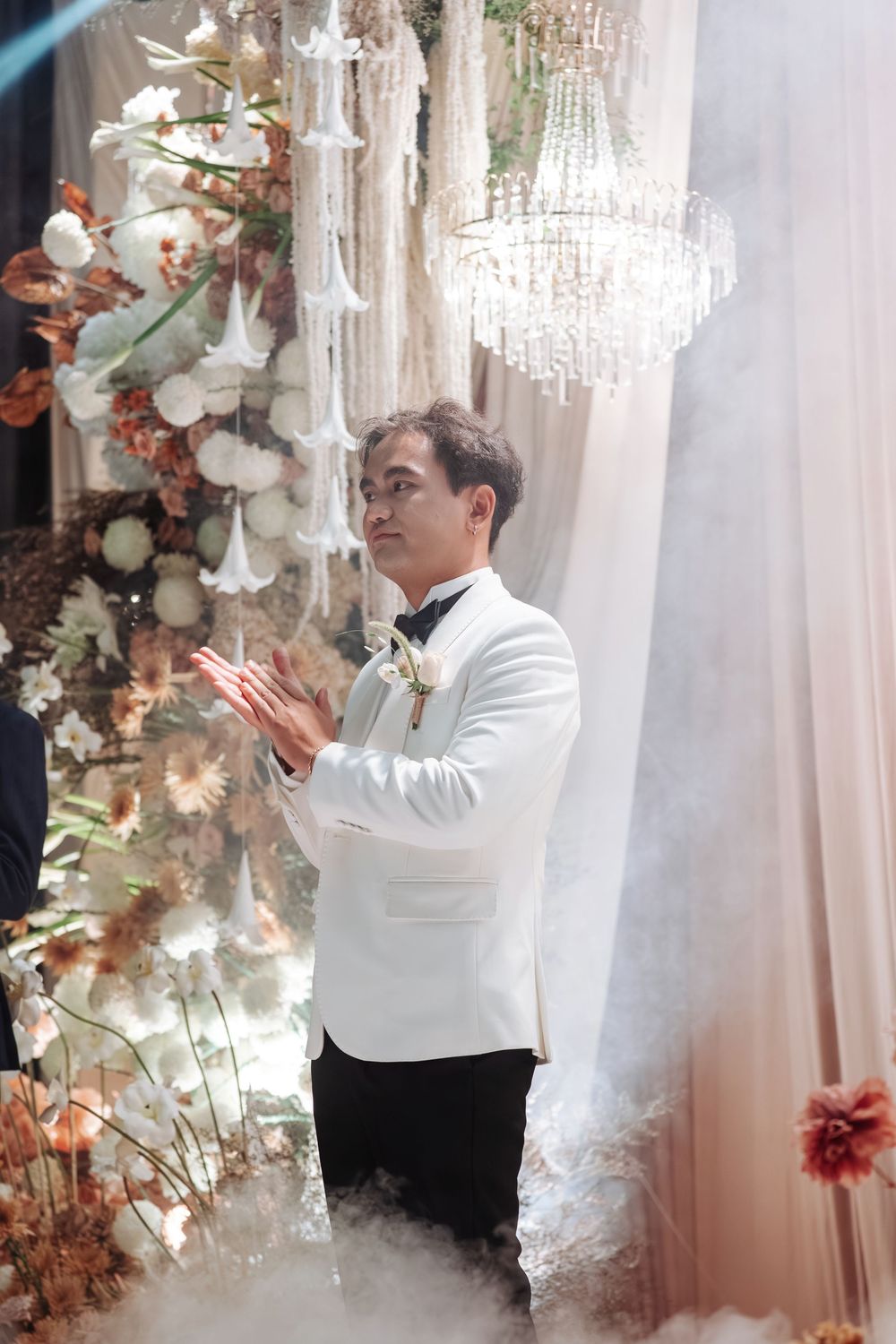Groom in white tuxedo clapping amid fog and floral decor at elegant wedding reception.