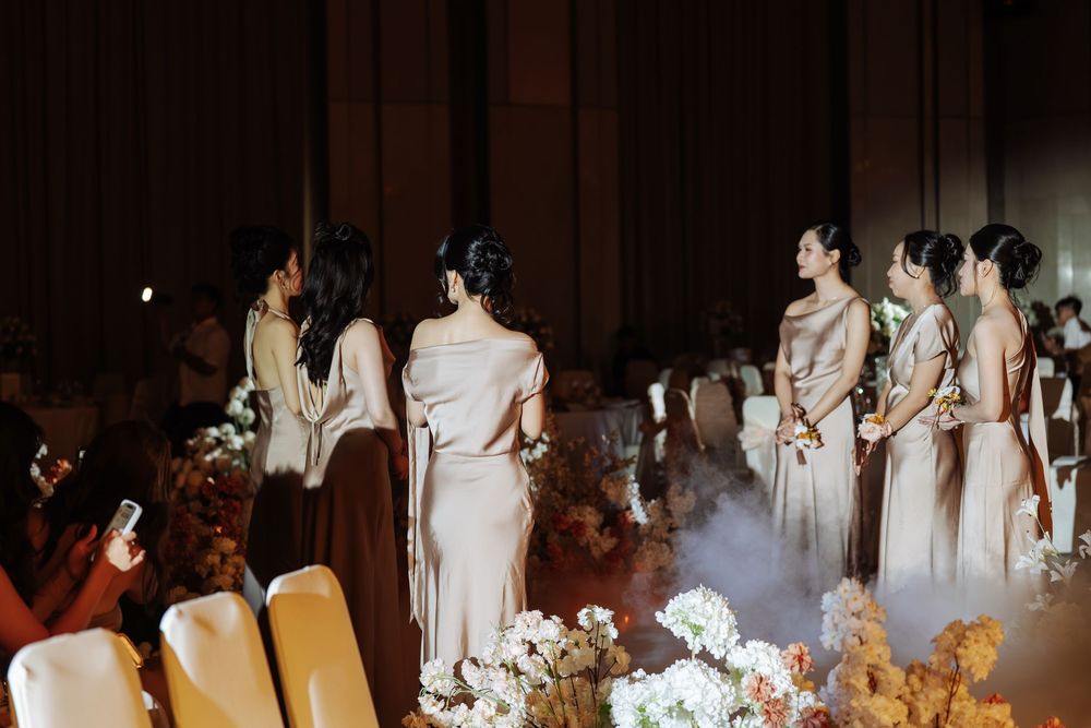 Bridesmaids in elegant taupe gowns stand in a row at a dimly lit wedding reception with floral arrangements.