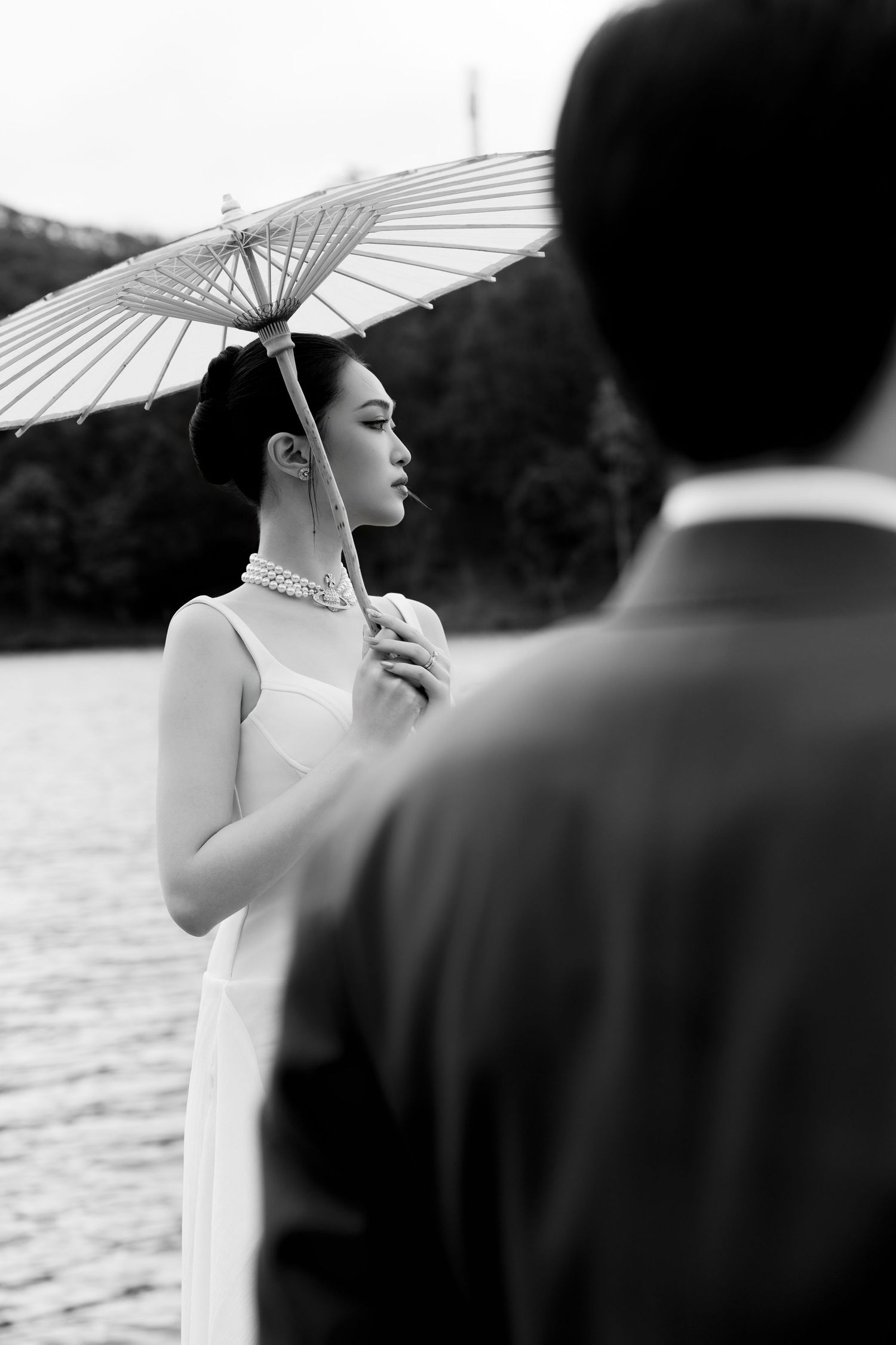 Black and white photo of elegant bride in white dress holding a parasol near a lake, groom in background.