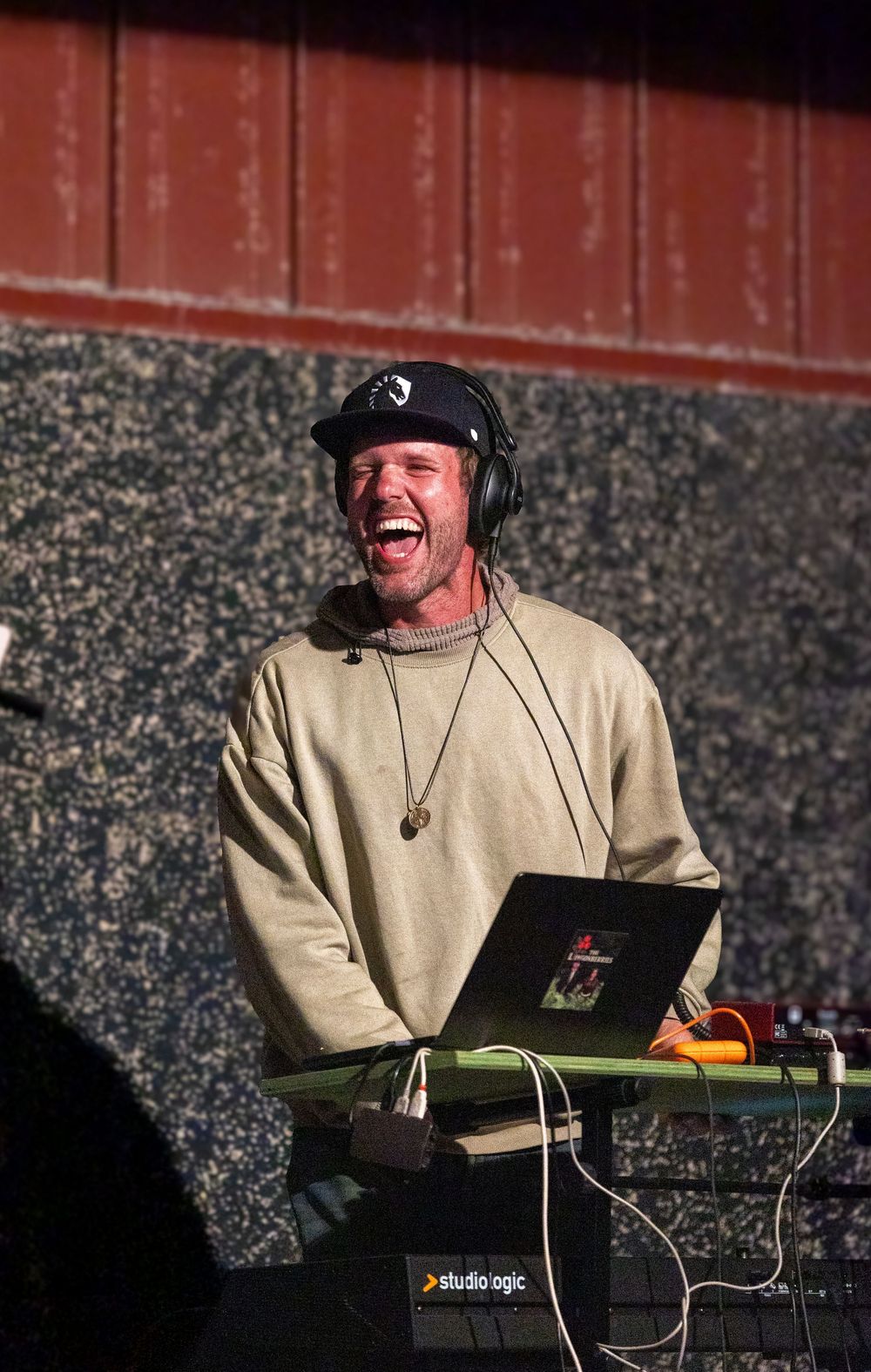 A DJ performs at an outdoor event wearing headphones and a beige sweater while working with equipment against a brick wall.