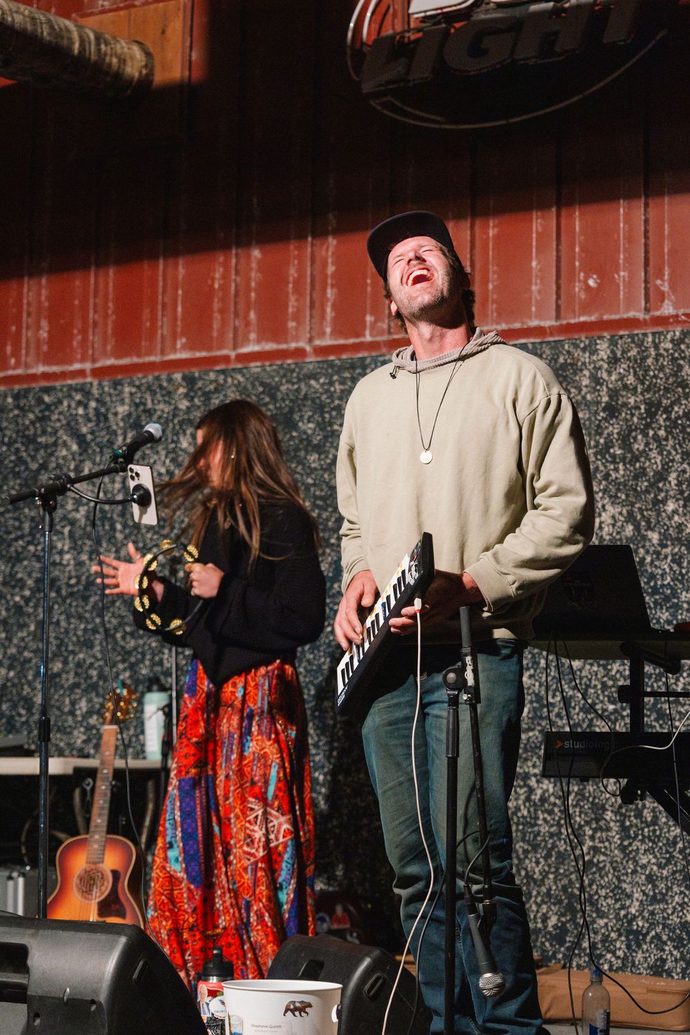 Musicians performing on stage with acoustic guitars against a patterned wall backdrop.