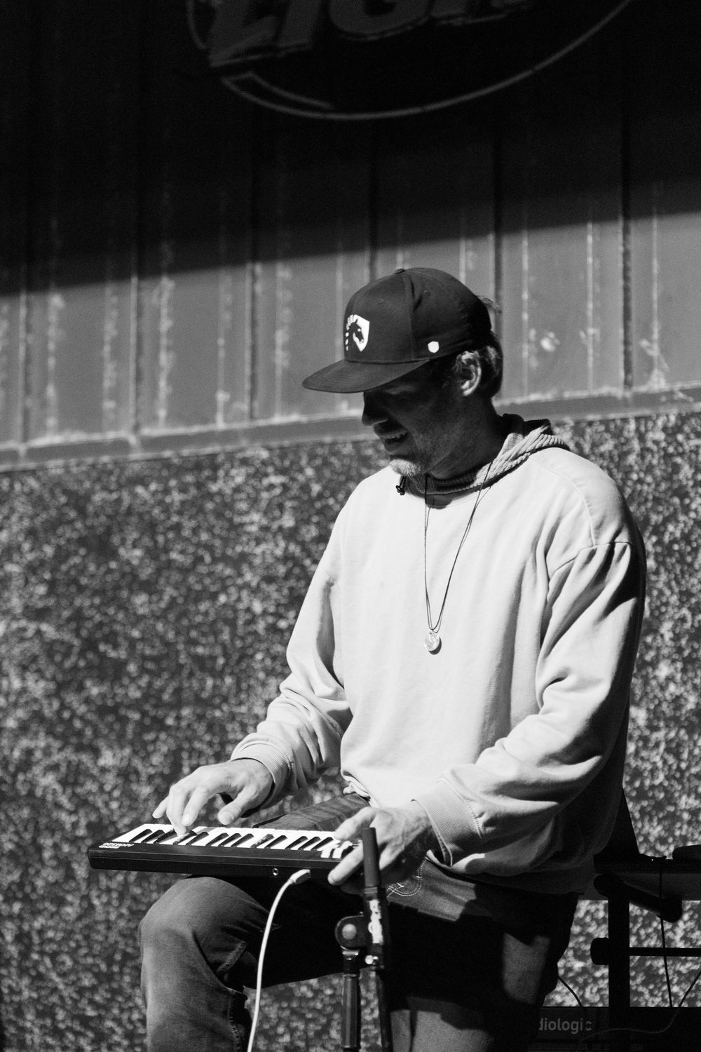 A musician in a baseball cap and hoodie plays a keyboard synthesizer outdoors in black and white.