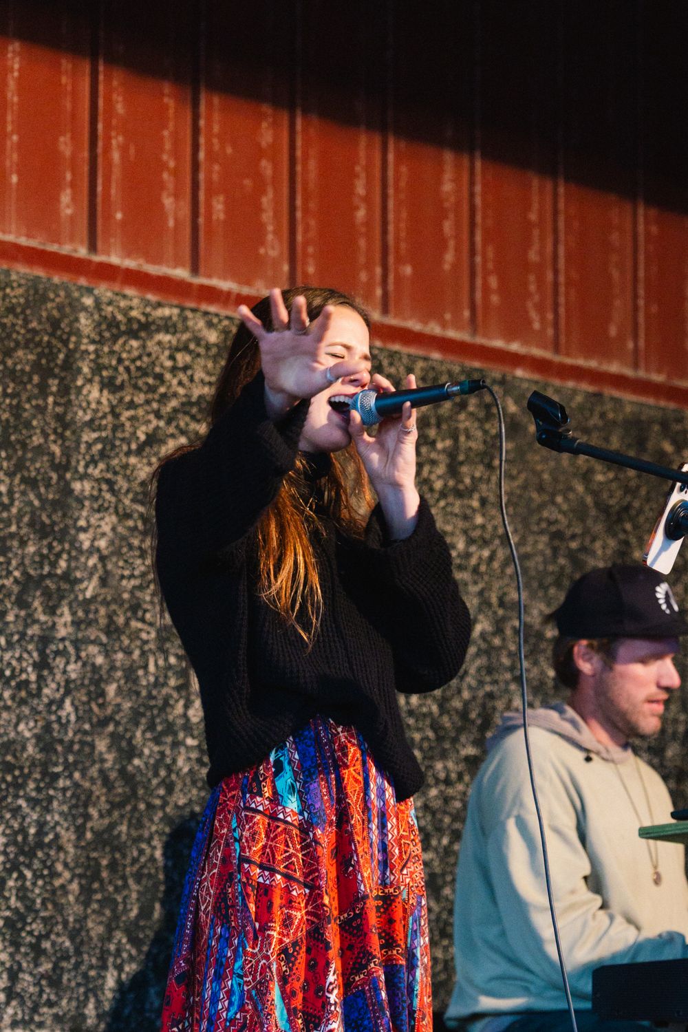 Someone performs with a microphone while wearing a colorful red and blue sequined vest against a textured wall.
