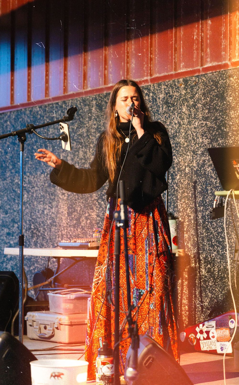 Performer in black top and orange patterned skirt passionately singing on stage with dramatic lighting and sparkly backdrop.