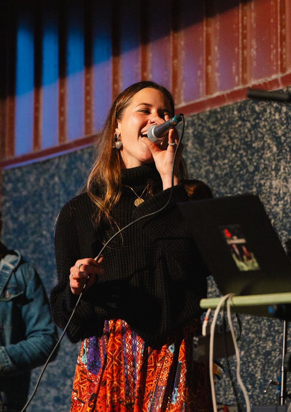 Singer performing emotional vocals in a black sweater with decorative skirt against a blue and red lit stage.