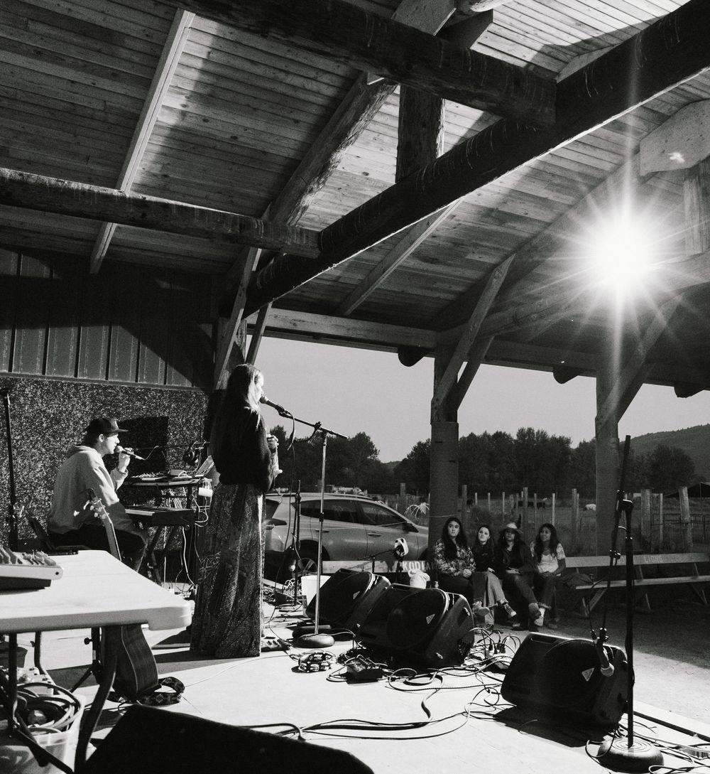 Musicians perform on an outdoor covered stage with equipment and instruments set up under wooden beams at sunset.