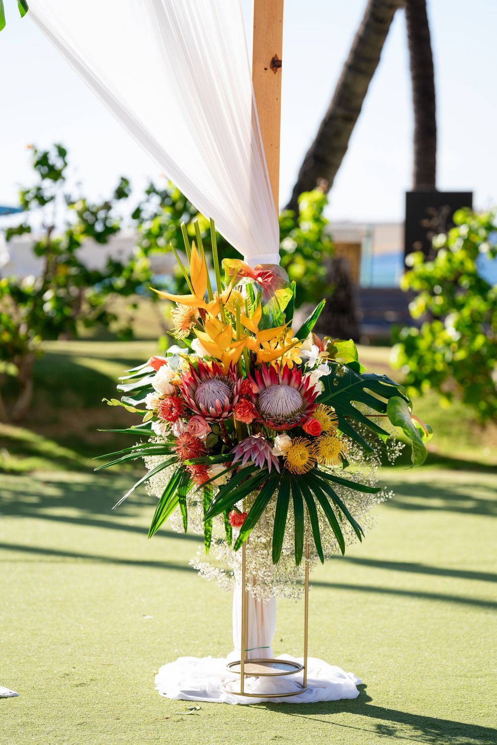 Three tropical floral arrangements with orange lilies and red protea flowers decorate a grassy outdoor wedding venue.