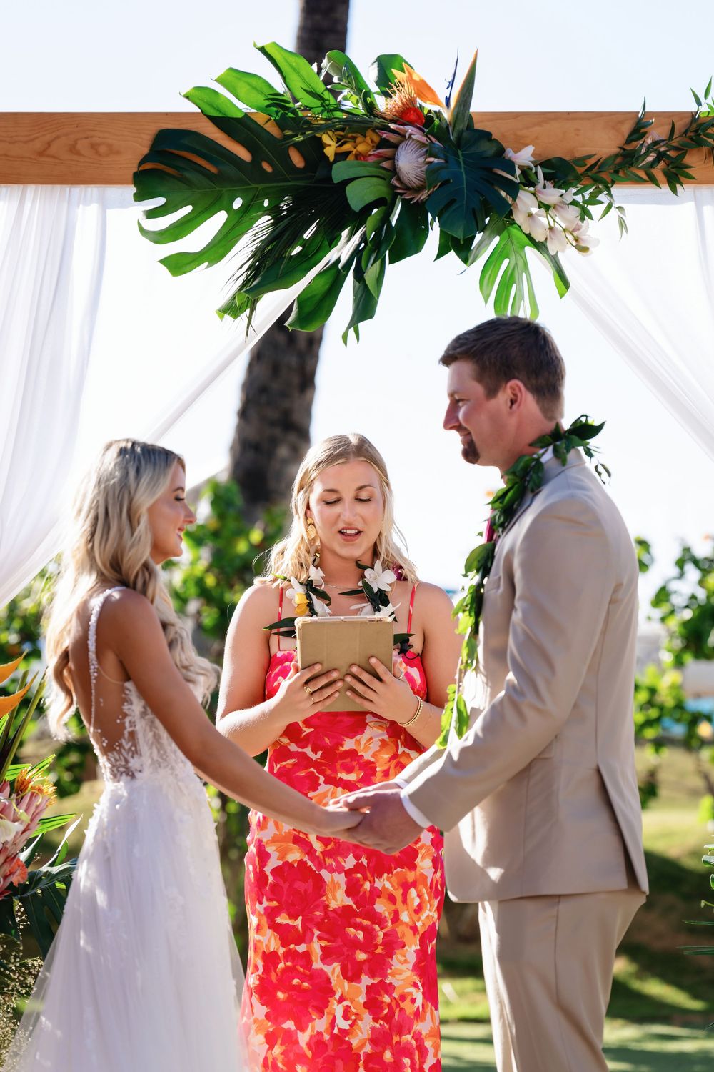 Wedding party members share emotional embraces during an outdoor tropical ceremony with palm trees and white drapes.