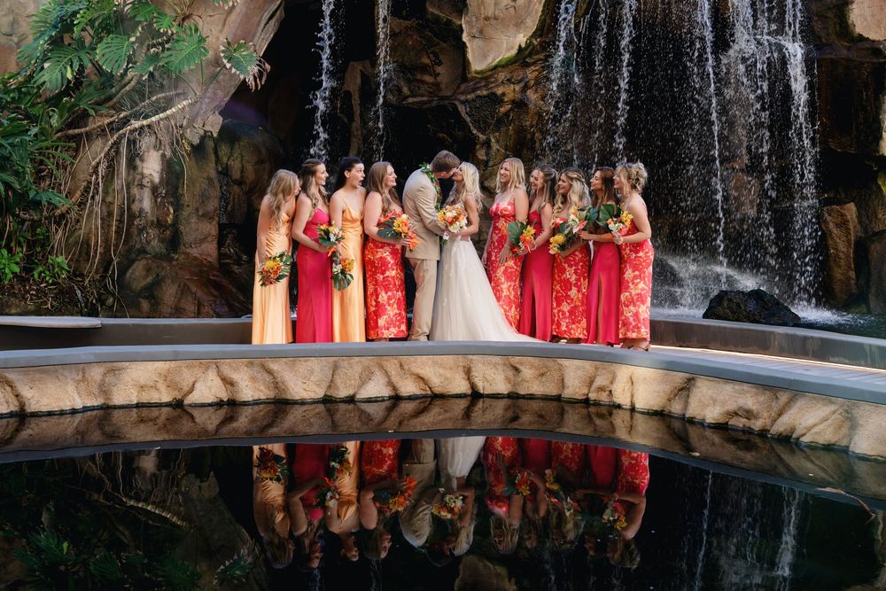 Wedding party in red and champagne dresses poses in front of waterfall with reflection in pool below.