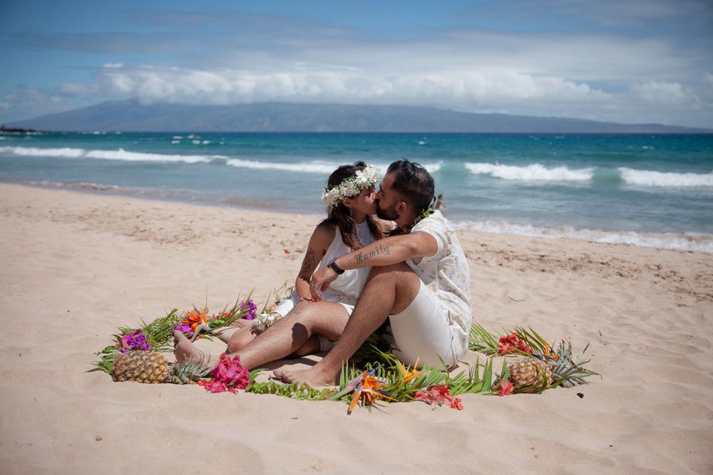 Couple lounging on sandy beach surrounded by colorful tropical flowers with ocean waves in background.