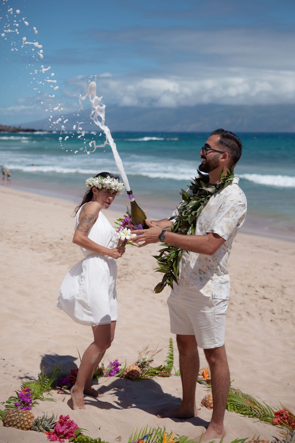 A celebratory beach moment captured in sequence as champagne sprays across a tropical Hawaiian shoreline with flowers in the sand.