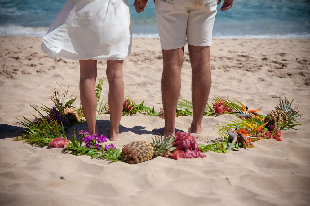 Multiple pairs of feet standing on tropical beach sand with pineapple and floral lei decorations below them.