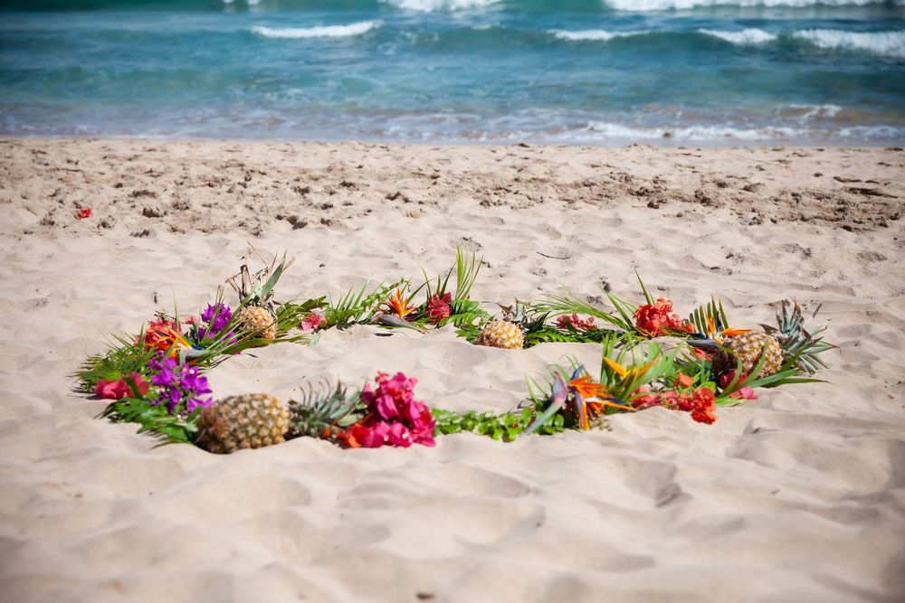 A heart shape made from tropical flowers and greenery arranged on sandy beach with ocean waves in the background.